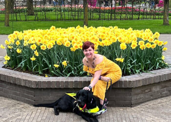A blind woman sits on brick wall and strokes her guide dog with yellow flowers behind her